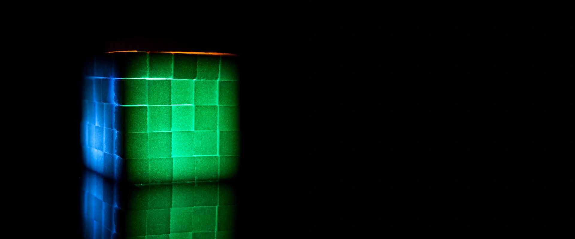 Sugar cubes stacked into a cube, lit green, blue and orange against black.